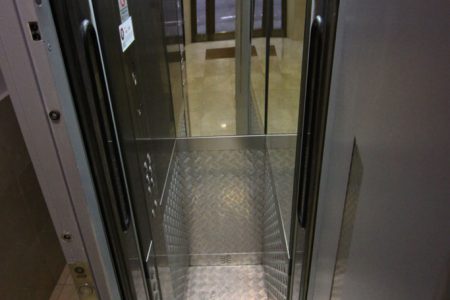 Puerta semiautomática abierta de un ascensor pequeño con cabina de puerta bus y chapa estriada