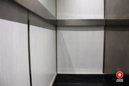 Interior de una cabina de ascensor con paredes de Formica y suelo negro, con zócalos inoxidables