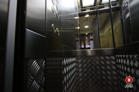 Interior de una cabina de ascensor con paredes de chapa estriada, medio espejo y botonera en columan